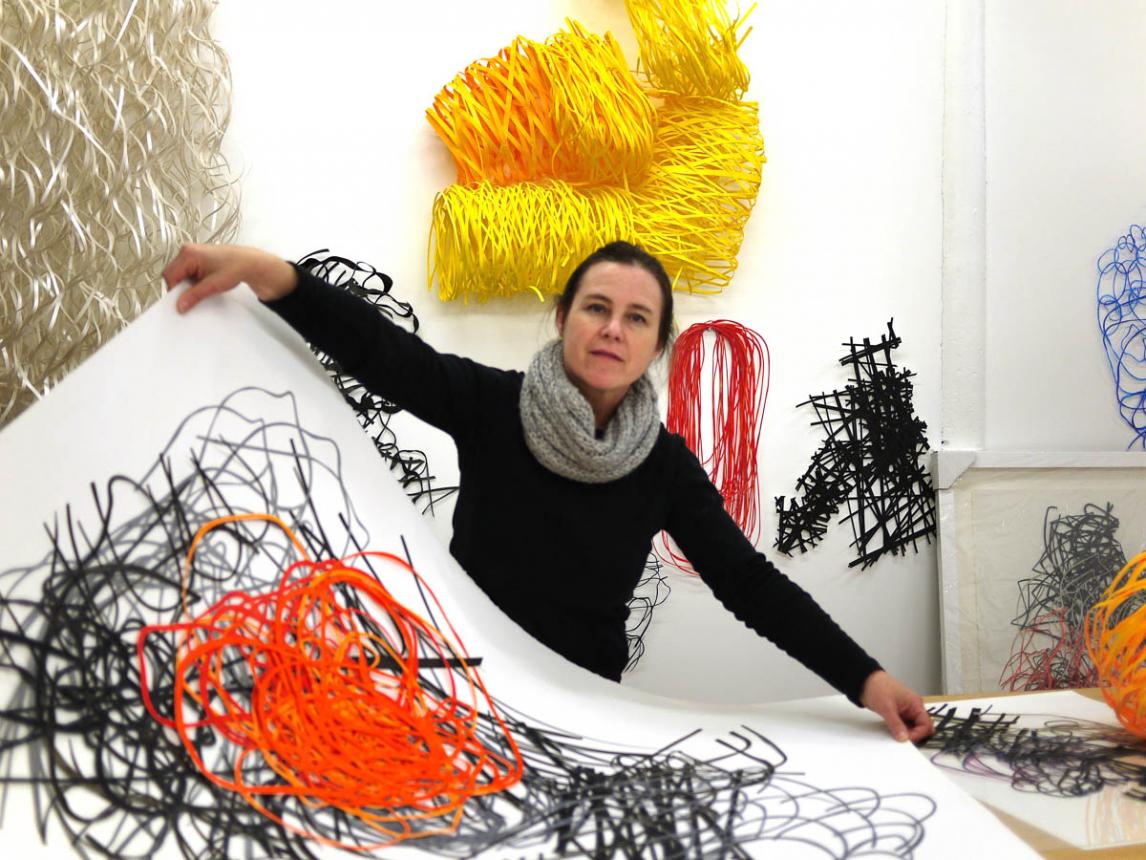 Artist Barbara Owens in her studio with many wall pieces around here and drawings on paper in front