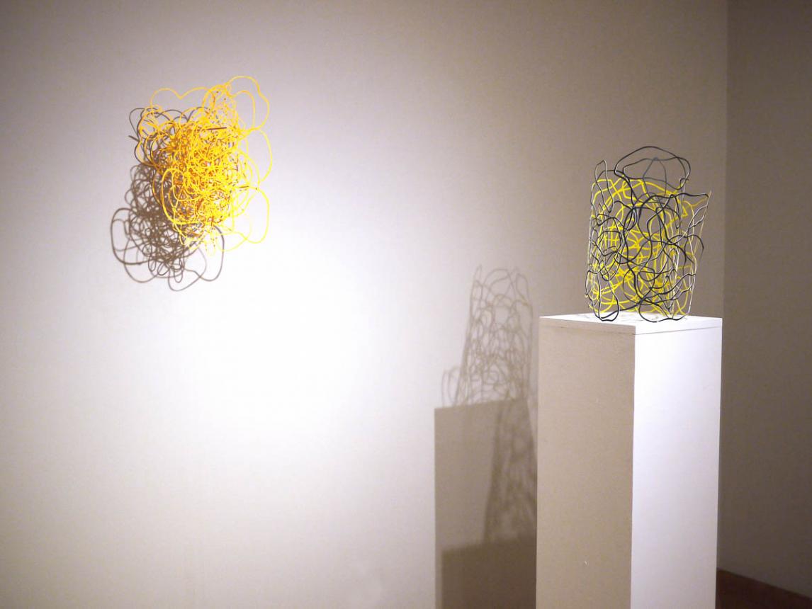 Gallery room view the yellow yarn wall piece on the wall and another yellow and black piece on a plinth in front